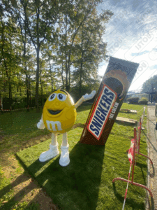 Une mascotte M&M accompagnée d’un chocolat Snickers, mise en scène dans un espace vert convivial et attrayant.