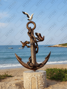 Impressionnante sculpture en bronze représentant une grande ancre marine entourée de chaînes et de trois mouettes en plein vol. L'œuvre repose sur un socle en pierre sur une plage ensoleillée, avec la mer d'un bleu profond et des vagues légères en arrière-plan. Deux petits bateaux de pêche sont visibles au large. Cette image est parfaite pour illustrer les thèmes de voyage, de la côte, de la pêche, et des symboles maritimes.