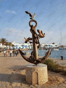 Impressionnante sculpture en bronze représentant une grande ancre marine entourée de chaînes et de trois mouettes en plein vol. L'œuvre repose sur un socle en pierre sur une plage ensoleillée, avec la mer d'un bleu profond et des vagues légères en arrière-plan. Deux petits bateaux de pêche sont visibles au large. Cette image est parfaite pour illustrer les thèmes de voyage, de la côte, de la pêche, et des symboles maritimes.
