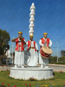 Sculpture représentant trois musiciens traditionnels tunisiens vêtus de costumes blancs et rouges, coiffés d'un chapeau jaune, jouant du 'Zokra' (hautbois), du 'Tabal' (tambour) et portant une pile de poteries ('Guerbou') décorées du drapeau tunisien. Ce monument célèbre la culture et la musique populaire tunisiennes, parfait pour un article sur les traditions ou les arts du spectacle