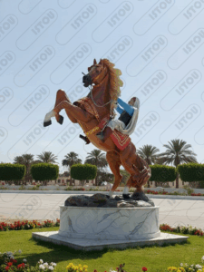 Statue colorée d'un cavalier en tenue traditionnelle montant un cheval brun cabré (s'élevant sur ses pattes arrière) sur un socle en marbre. Le décor autour du monument est paysager avec des palmiers et des fleurs. Cette œuvre représente le patrimoine équestre et la culture locale, idéale pour des articles sur les traditions, l'histoire ou les sites touristiques du Maghreb.