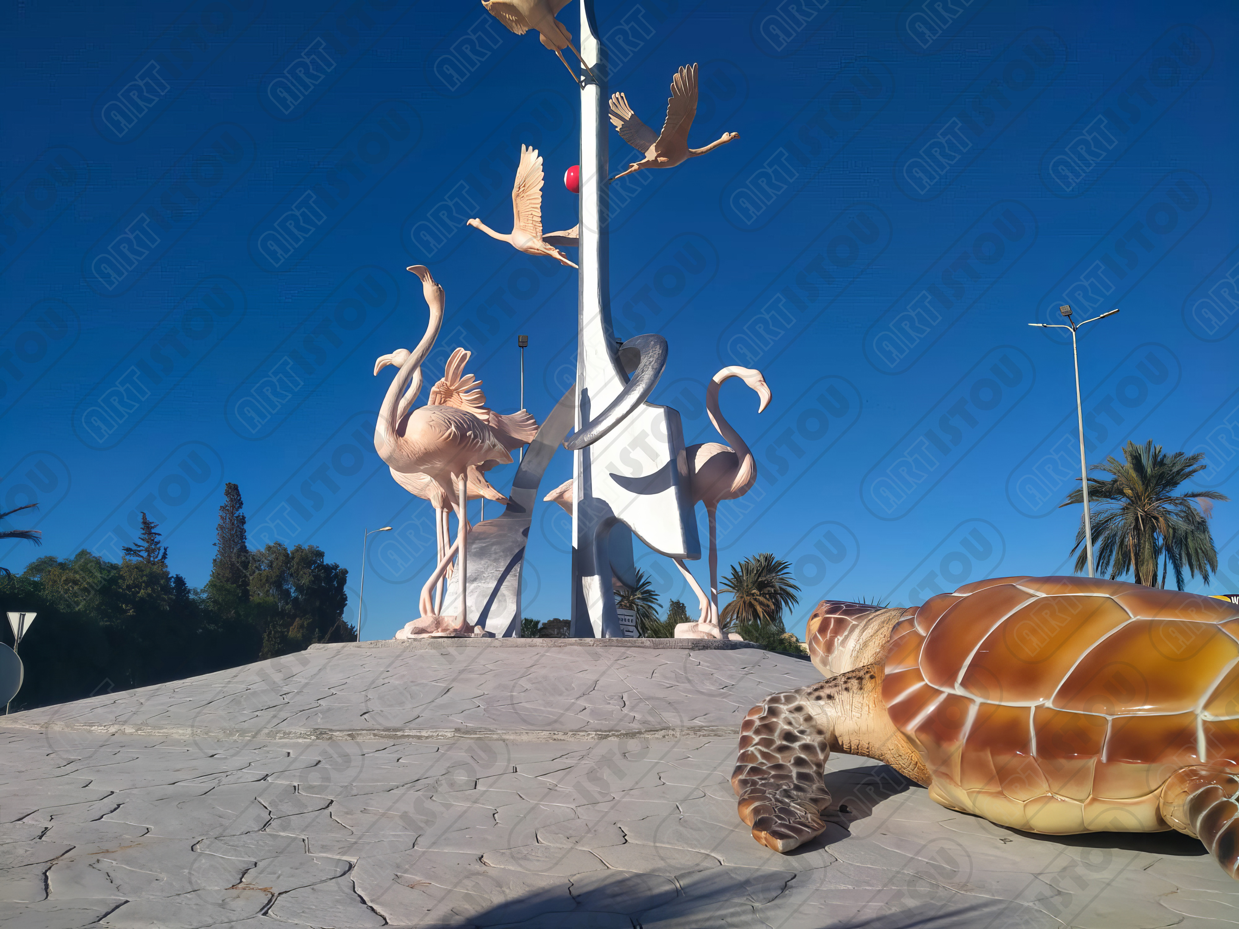 Monument sur un rond-point représentant la faune locale. Au centre, deux grands flamants roses (flamingos) et des oiseaux en vol au sommet. Au premier plan, une grande sculpture réaliste d'une tortue de mer. L'œuvre symbolise la biodiversité et la protection des espèces marines et aviaires de la région méditerranéenne (Tunisie). Parfait pour les articles sur l'écotourisme ou les réserves naturelles.