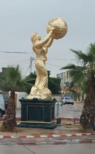 Impressionnante statue dorée d'une femme élancée, debout sur un socle de marbre vert, levant un globe terrestre doré au-dessus de sa tête. Entourée de palmiers dans un environnement urbain, cette sculpture représente probablement la prospérité, l'espoir ou la vision mondiale. Idéale pour des articles sur l'urbanisme, l'art public ou les symboles locaux.