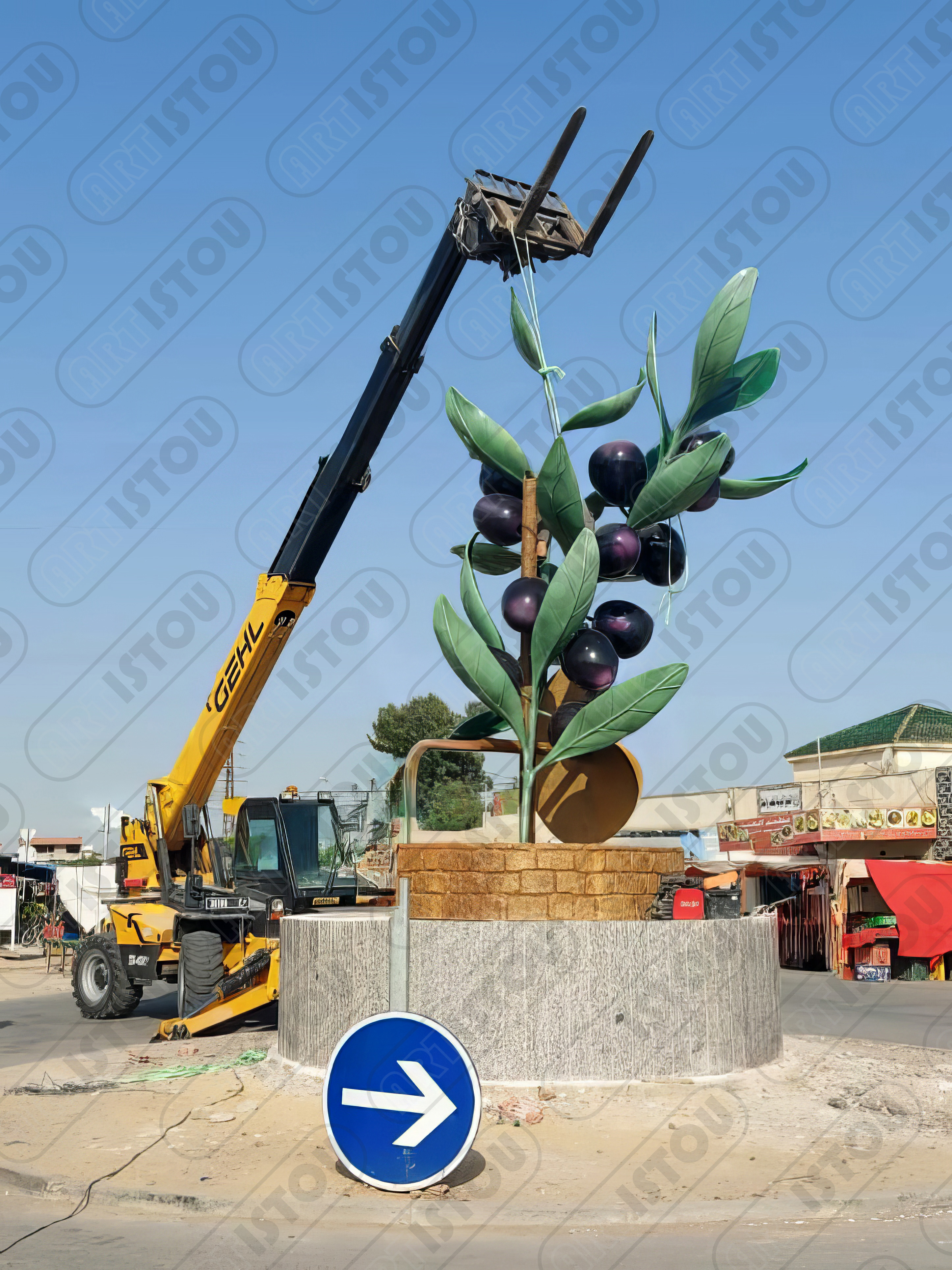 Sculpture monumentale en cours d'installation, montrant un rameau d'olivier géant avec des olives noires proéminentes, près d'une grenade (fruit) au sommet d'un puits stylisé. L'œuvre repose sur un socle en pierre et célèbre l'agriculture, le patrimoine oléicole et les produits emblématiques de la région.